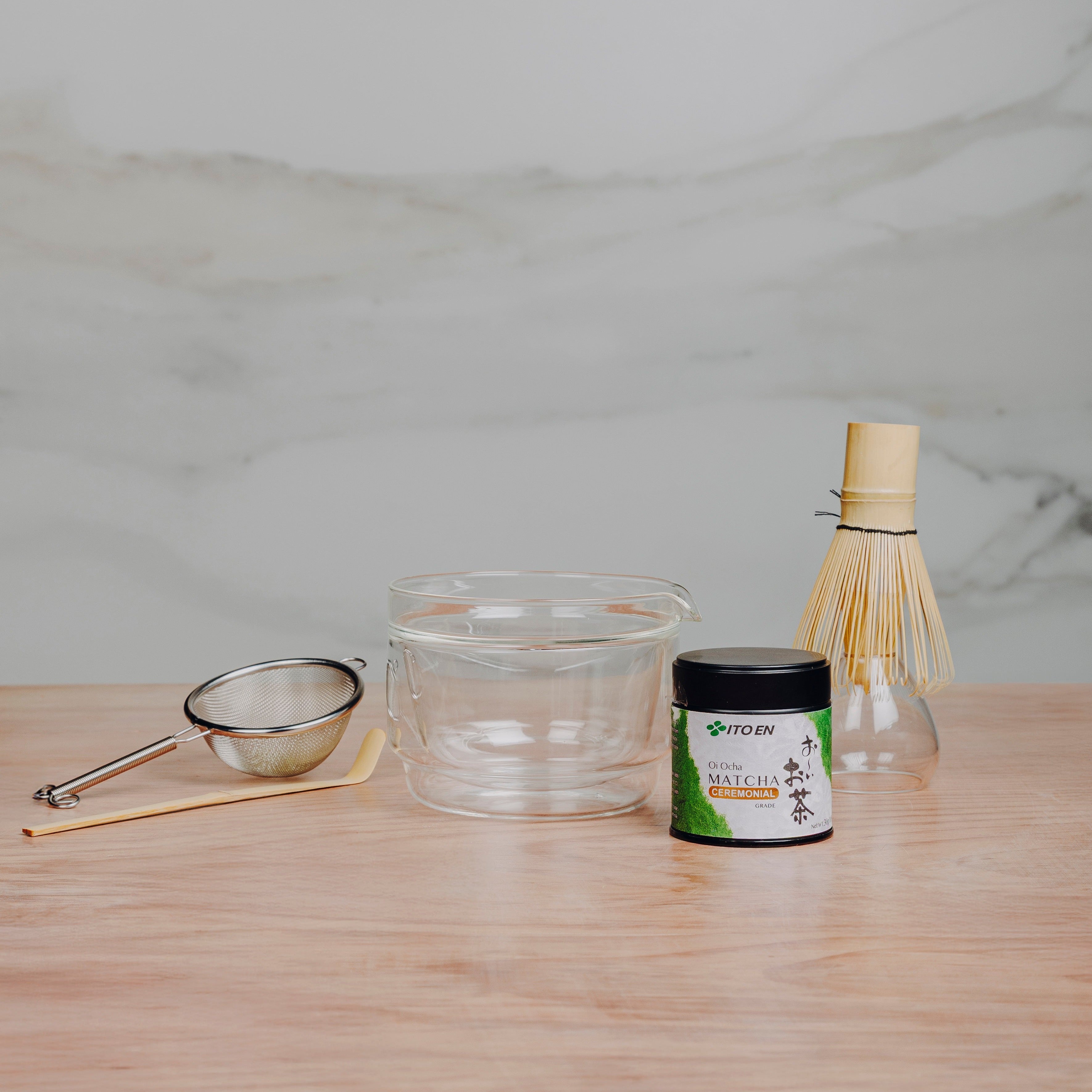 Tea-making tools including a whisk, sieve, and container on a wooden surface with a marble background.